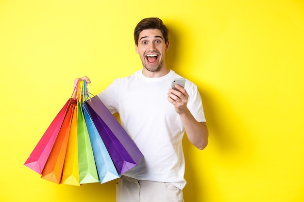 Homem feliz recebe cashback pela compra, segurando um smartphone e sacolas de compras, sorrindo animado, em pé sobre a parede amarela