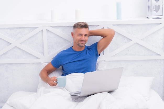 Homem feliz relaxa tomando café e usando o laptop na cama de manhã, relaxamento