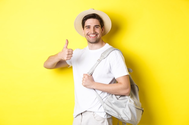 Homem feliz saindo de férias, segurando a mochila e mostrando o polegar, sorrindo satisfeito, encostado na parede amarela