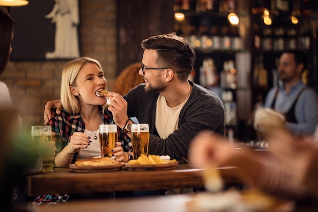 Homem feliz se divertindo com sua namorada e alimentando-a enquanto bebe cerveja em um pub