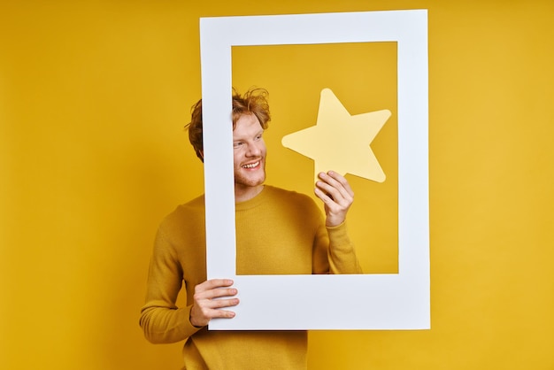 Homem feliz segurando estrela enquanto olha através de um porta-retrato contra fundo amarelo