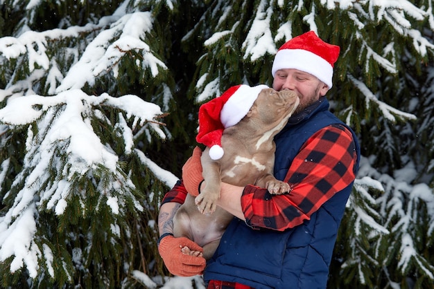 Homem feliz segurando lindo cachorro nas mãos usando um chapéu de Papai Noel na floresta de neve. Menino sorridente abraçando adorável cachorrinho em madeira de inverno. Amante de animais de estimação. Cão - conceito de amigo humano. Férias de natal