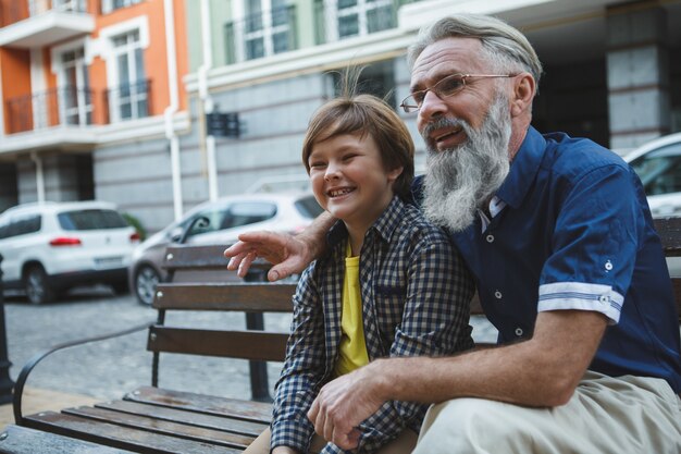 Homem feliz sênior abraçando seu neto sentados juntos no banco no centro da cidade