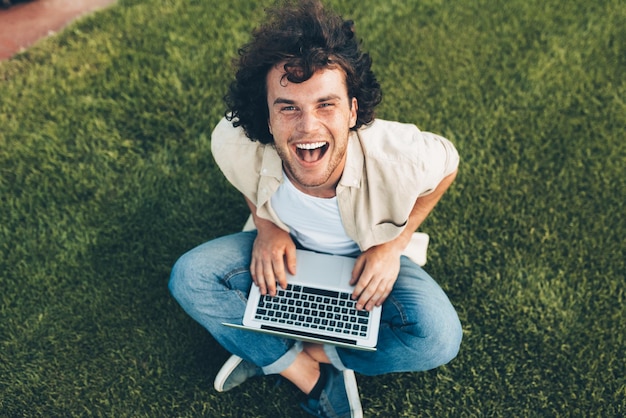 Homem feliz sentado no chão no parque trabalhando no laptop Homem sorridente com cabelos cacheados usando laptop para conversar online com amigos sentados na grama ao ar livre Empresários de tecnologia