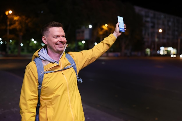 Homem feliz sorridente segurando smartphone e encontrando amigos ou chamando táxi no conceito de rua à noite