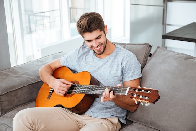 Homem feliz tocando violão enquanto está sentado no sofá em casa. Olhando para o violão.