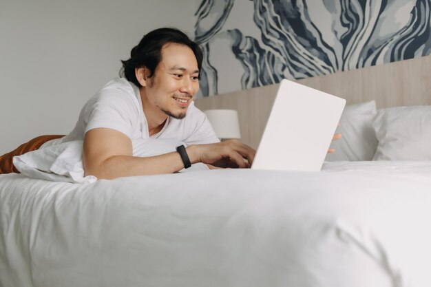 Homem feliz trabalhando com laptop na cama no conceito de trabalho do hotel