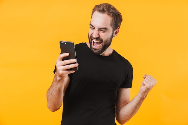 Homem feliz vestindo camiseta preta básica regozijando-se e segurando o smartphone isolado sobre a parede amarela