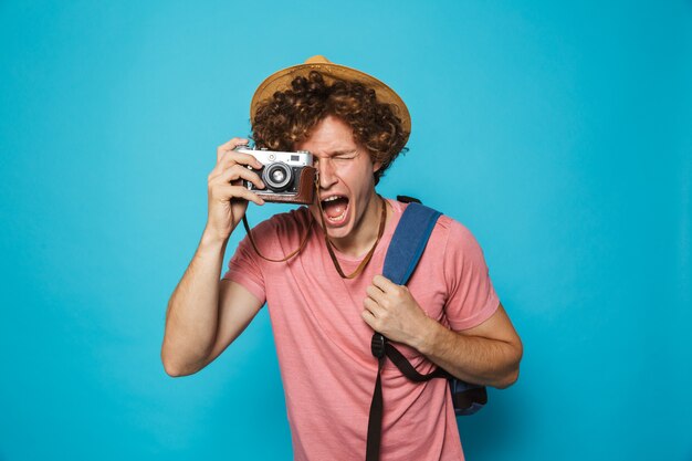 Homem feliz viajante com cabelos cacheados, usando mochila e chapéu de palha fotografando na câmera retro