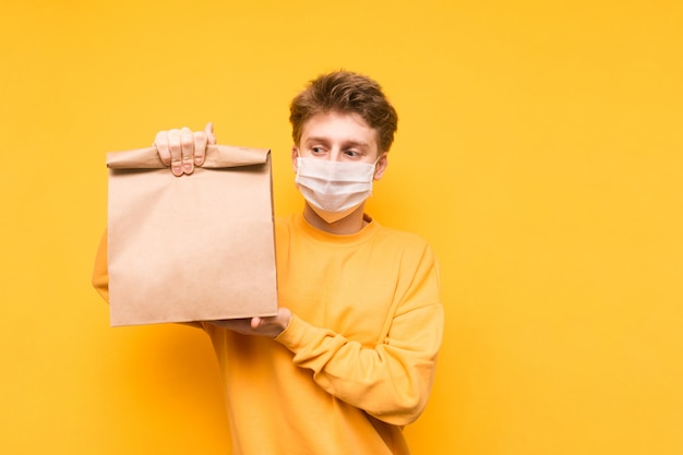 Homem fica com um pacote de comida da entrega em suas mãos em uma máscara médica no rosto, olha para a câmera e mostra um gesto bem.