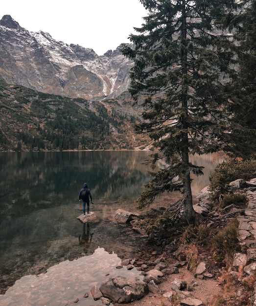 Homem fica em pé na grande pedra no meio de um lago cercado por altas montanhas