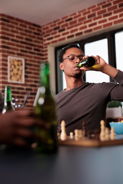 Homem focado bebendo bebida alcoólica enquanto pensa no próximo movimento de xadrez. pessoa inteligente bebendo cerveja com amigos enquanto relaxa em casa na sala de estar com jogo de xadrez e lanches.