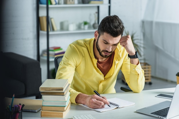 homem focado escrevendo em caderno perto de laptop e livros em escritório moderno