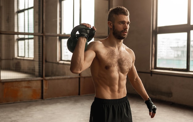 Homem forte e confiante com barba e torso musculoso nu, fazendo exercícios de bíceps com kettlebell pesado durante um treino funcional intenso na academia