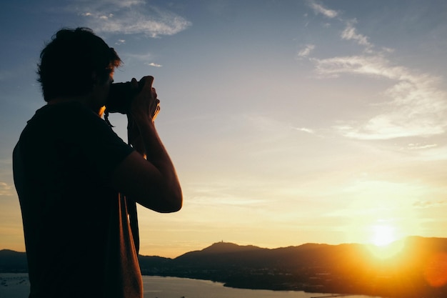 Homem fotografando enquanto está de pé contra o céu durante o pôr do sol