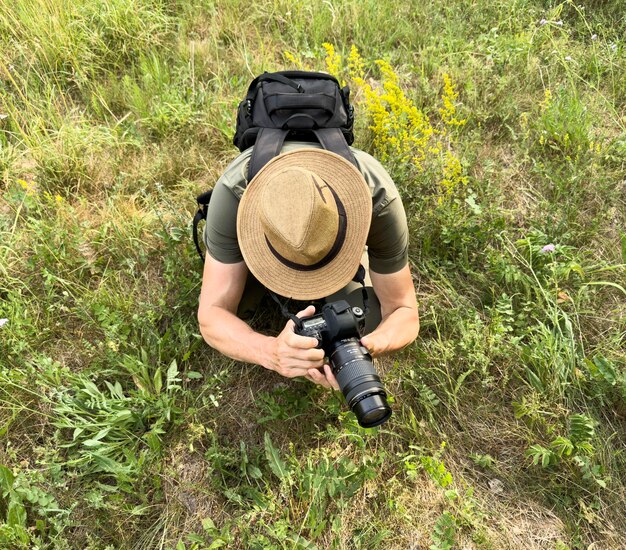 Homem fotógrafo de chapéu tirando foto na natureza entre grama verde campo selvagem segurando câmera DSLR na mão