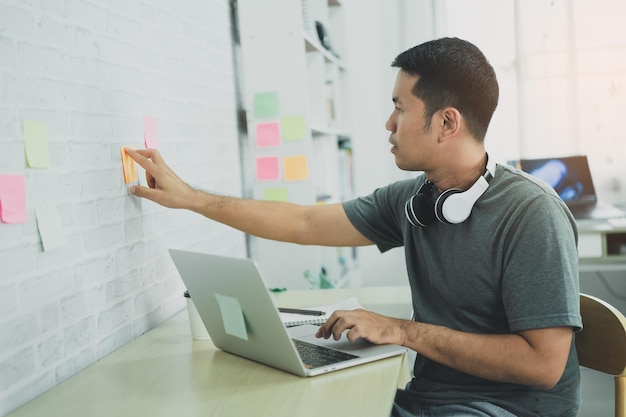 Homem freelance asiático sorrindo colar postit na parede para gravar para o trabalho e usar no laptop na mesa de madeira em casa Homem empresário trabalhando para seu negócio em casa Conceito de negócio