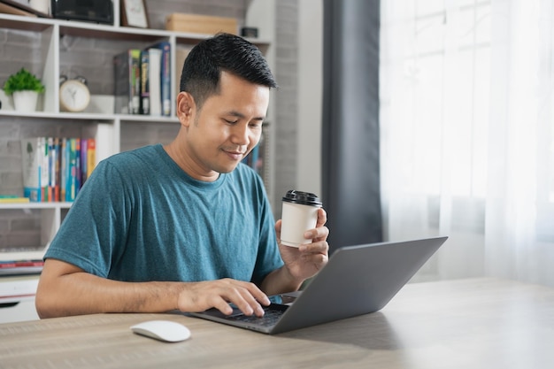 Homem freelance asiático sorrindo segurando a xícara de café quente e trabalhando no computador portátil na mesa de madeira em casa Homem empresário trabalhando para seu negócio em casa Trabalho de negócios em casa conceito