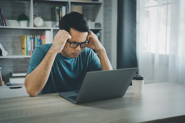 Homem freelance asiático usa óculos e passa a cabeça tendo um tempo triste de depressão estressante enquanto trabalhava no laptop em casa Homem de depressão triste serios trabalhando em casa Trabalhando em qualquer lugar conceito
