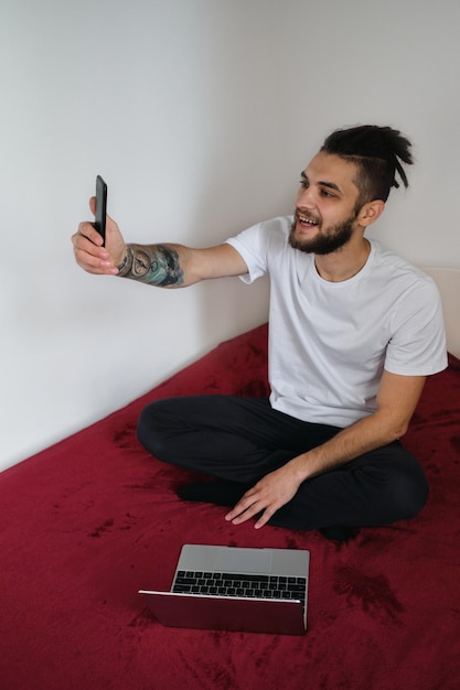 Homem freelancer bonito em camiseta branca com barba e dreadlocks trabalha em casa em auto-isolamento