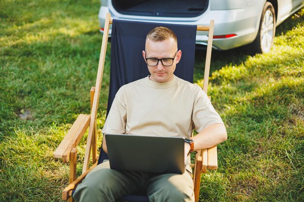 Homem freelancer senta-se em uma cadeira de madeira na natureza e trabalha online em um laptop Um homem viaja e trabalha remotamente em um laptop Trabalho de escritório na natureza Férias