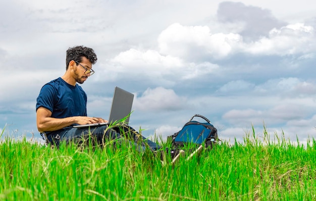 Homem freelancer sentado com laptop e mochila no campo Homem sentado no campo verde trabalhando em seu laptop Conceito de homem freelancer trabalhando em campo