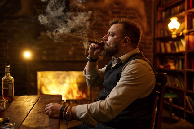 Foto homem fuma um charuto e bebe bebida alcoólica, uma estante de livros e um rico interior de escritório no fundo