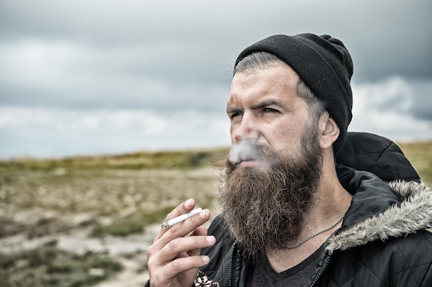 Homem fumando cigarro, jovem hipster barbudo de chapéu e jaqueta na montanha e céu nublado em fundo natural, copie o espaço