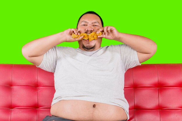 Homem ganancioso com sobrepeso comendo comida lixo de frango frito enquanto está sentado no sofá isolado em fundo verde