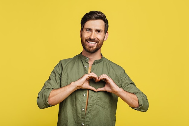 Homem gentil moldando as mãos como coração fazendo gesto de amor perto do peito expressando bondade e sorrindo para a câmera