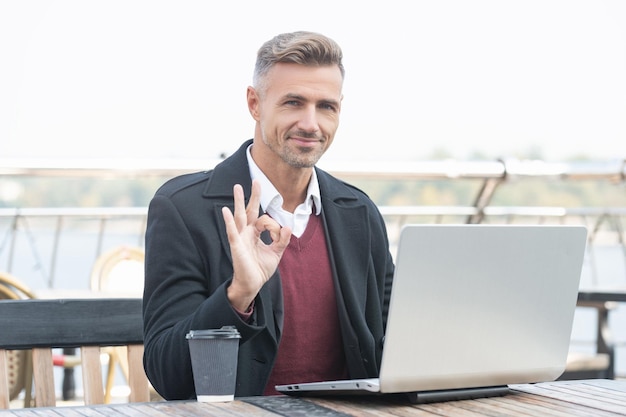 Homem gerente feliz mostra gesto de anel bem fazendo sinal de OK trabalhando no laptop no café ao ar livre