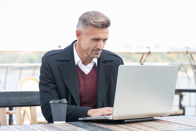 Homem gerente sério trabalhando online no laptop tomando café no café ao ar livre