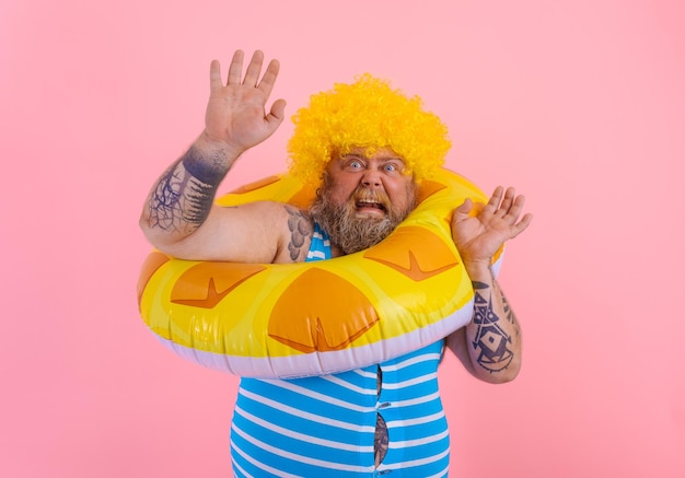 Homem gordo com medo com peruca na cabeça está pronto para nadar com um salva-vidas de rosquinha