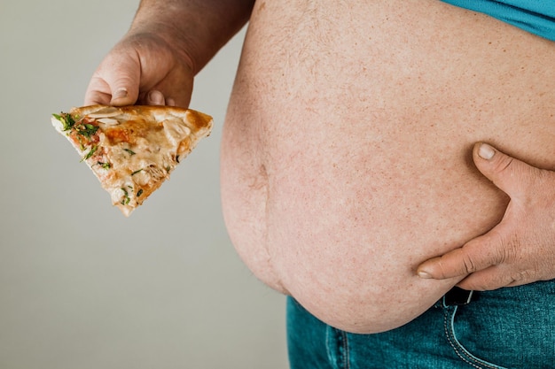 Homem gordo com uma fatia de pizza em um fundo cinza o conceito de alimentos não saudáveis