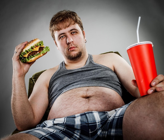 Homem gordo comendo hambúrguer sentado na poltrona. Estilo fast-food.