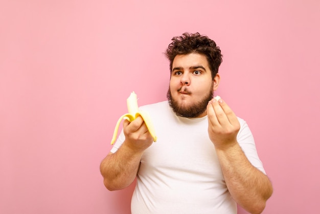 Homem gordo encaracolado engraçado com barba come uma banana em um fundo rosa desvia o olhar com uma cara engraçada