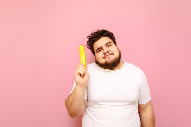 Homem gordo engraçado fica em um fundo rosa com uma banana na mão olha na câmera
