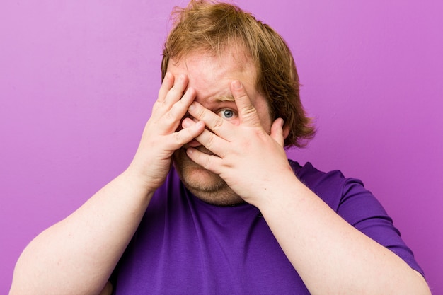 Homem gordo jovem ruiva autêntica pisca entre os dedos, assustado e nervoso.