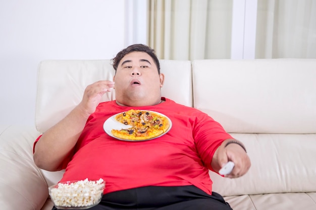 Homem gordo mascando pipoca enquanto segura o controle remoto da tv