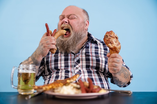 Foto homem grande e faminto a comer salsichas à mesa com comida rica e uma caneca de cerveja