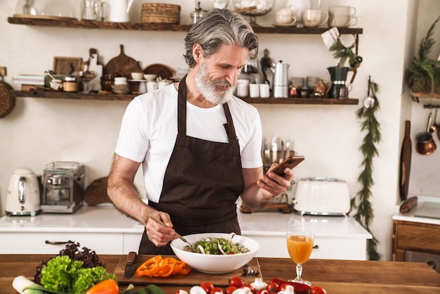 Foto homem grisalho de avental satisfeito usando o celular e preparando salada para o almoço em uma cozinha aconchegante