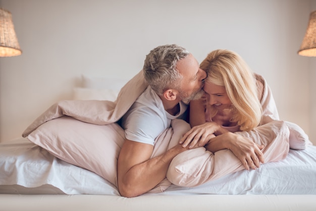 Homem grisalho e uma mulher loira deitados na cama com ar romântico