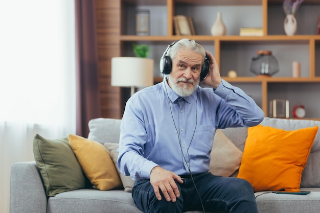 Homem grisalho sênior sentado no sofá em casa relaxando e ouvindo música em fones de ouvido usando o aplicativo de música no telefone