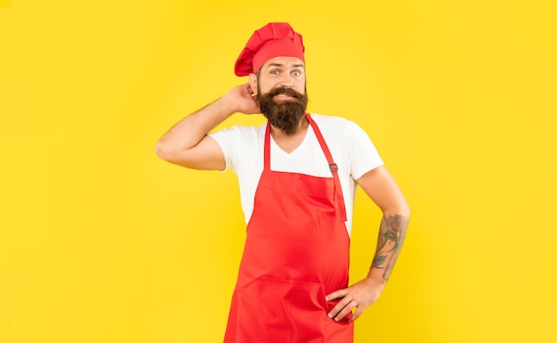 Homem hesitante de touca vermelha e avental coçando a cabeça cozinheiro de fundo amarelo