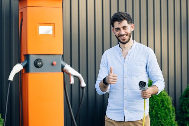 Homem hipster barbudo caucasiano parado perto da estação de carregamento elétrico olhando para a câmera