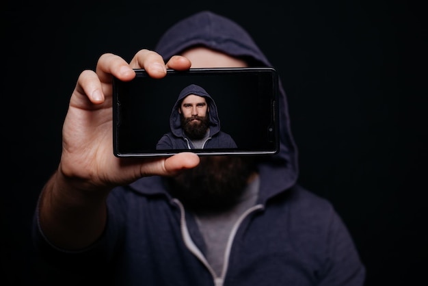 Foto homem hipster com barba tirando foto de self-portrait de smartphone visualização de tela estúdio de instantâneo em um fundo preto