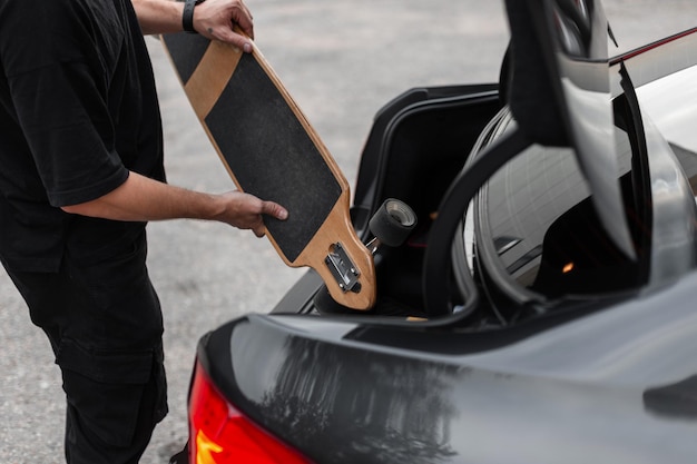 Homem hipster em roupas pretas coloca um longboard no porta-malas do carro Fins de semana ativos e descanso