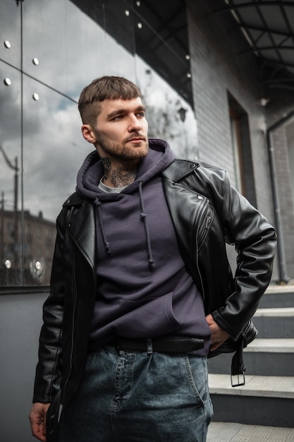 Homem hipster modelo bonito de moda bem-sucedida em roupas casuais da moda com uma jaqueta de couro rock e capuz andando na cidade