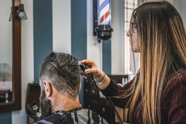 Homem hipster no salão de barbearia cortando barba e cabelo Mulher de cabeleireiro usando máquina de cortar cabelo para corte moderno com desenho geométrico na nuca Conceito de barbearia