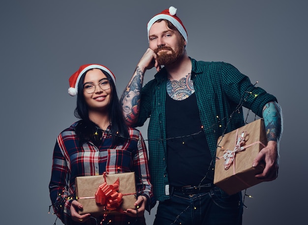 Homem hipster tatuado barbudo e mulher morena sexy vestida com uma camisa de lã detém presentes de natal sobre fundo cinza.